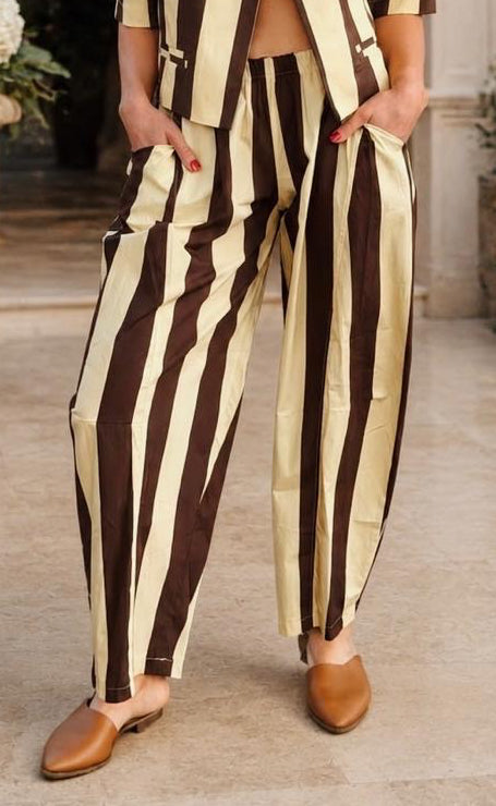 A woman modeling a matching two-piece striped suit in a beige and dark brown colorway, standing in an elegant outdoor courtyard with stone arches and potted plants.